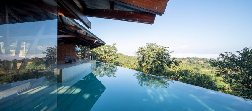 A home with glass walls next to an infinity pool overlooking treetops