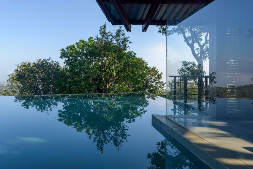 A home with glass walls next to infinity swimming pool overlooking treetops