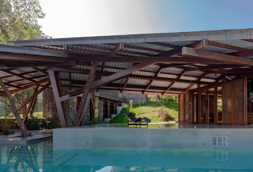 A terrace with a swimming pool with an angular metal roof supported by a wooden structure