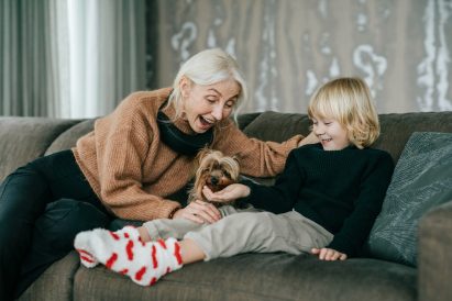 Photo of a woman wearing a black air purifier around her neck while patting a small dog and playing with a child on a sofa