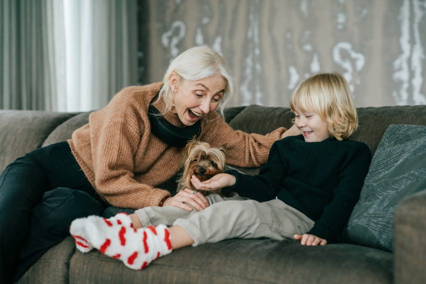Photo of a woman wearing a black air purifier around her neck while patting a small dog and playing with a child on a sofa