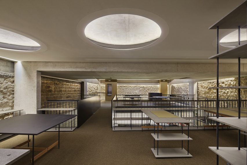 A room with dark carpets, circular recessed ceiling lights and furnished with tables and shelving