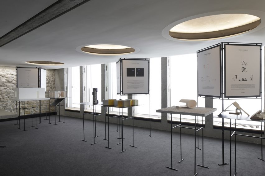 A room with grey flooring, white brick walls, large windows, circular recessed ceiling lights and freestanding displays