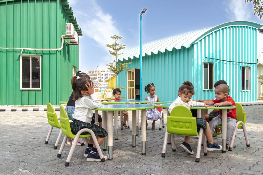Children sitting at outdoor tables and chairs with blue and green metals buildings behind