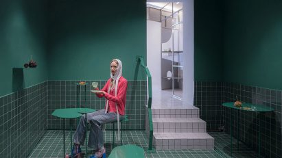 A dark green room with green floor tiles, curved green metal tables and a person sitting with a coffee and croissant