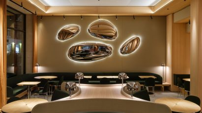 A restaurant interior with wood columns and beams, wood tables, green upholstered seating and oval metallic wall lights