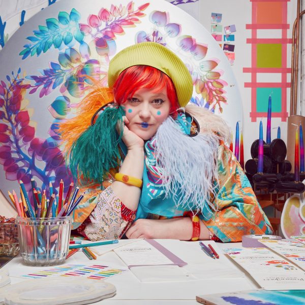 Person with red hair and green beret sitting at a table with colouring pencils