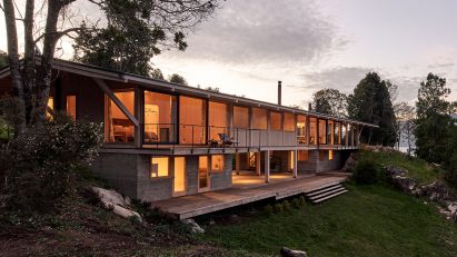 Timber house on a hillside with large windows revealing warmly-lit rooms