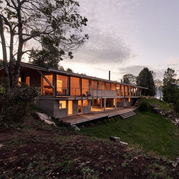 Timber house on a hillside with large windows revealing warmly-lit rooms