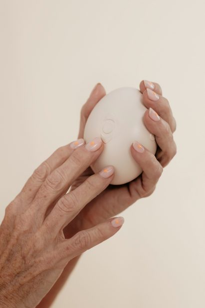 Photo of the hands of a middle-aged woman with multicoloured nail polish holding an oval-shaped off-white massager