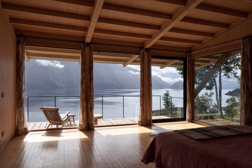 A bedroom in a timber structure with glass doors leading to an terrace overlooking a lake
