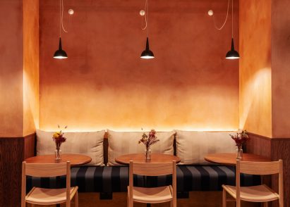 Restaurant interior with an orange ombre wall, black pendant lights, bench seating and wooden tables and chairs