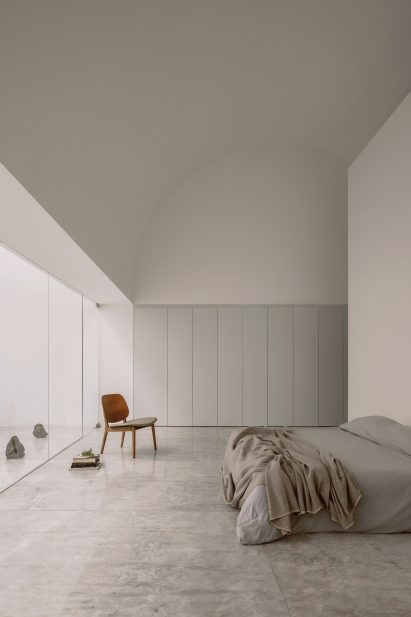 White interior room with an arched ceiling, built-in white wardrobes, a bed with grey bedding, a wooden chair and sliding glass doors