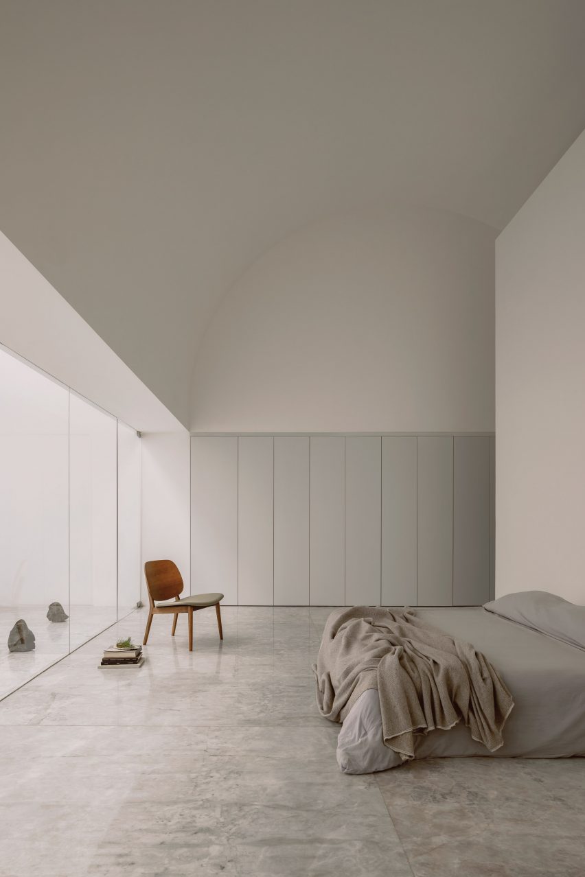 White interior room with an arched ceiling, built-in white wardrobes, a bed with grey bedding, a wooden chair and sliding glass doors