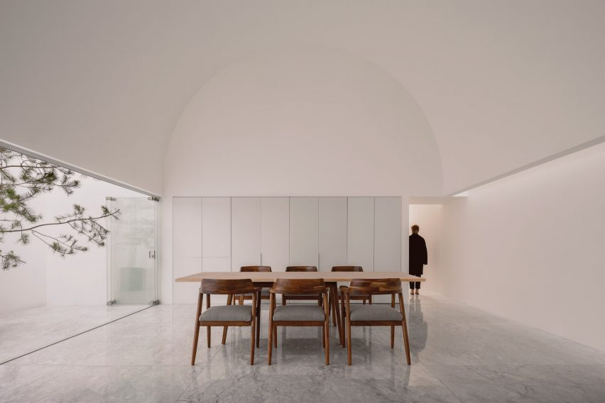 A white dining room with arched ceiling, wooden dining table and chairs, built-in white wall storage and sliding glass doors opening to a courtyard