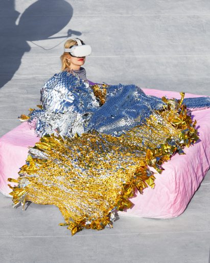 Model sitting on mattress wearing and covered in shiny metallic materials
