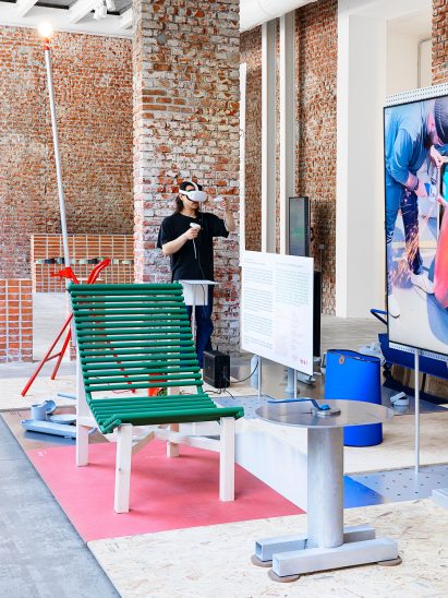 A recliner lounge chair made from a bathroom radiator painted green and laid over a wooden frame sits among other objects in an exhibition of objects made from scrap metal