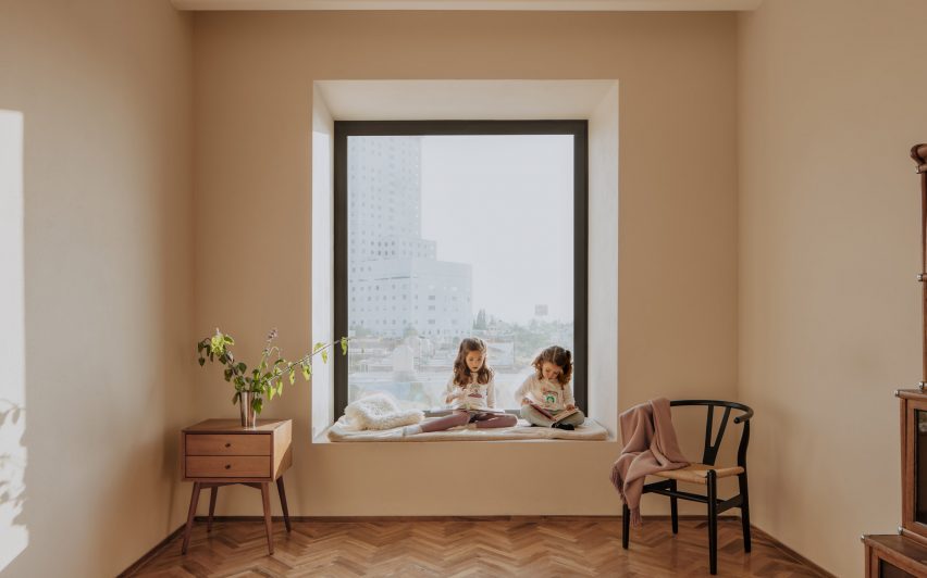 Internal room with white walls, wooden parquet flooring and children sitting in a square window