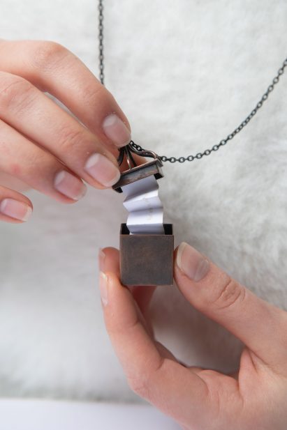 Person holding a small metal box on a necklace chain that opens to reveal folded paper