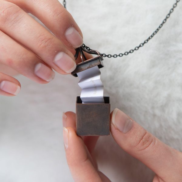 Person holding a small metal box on a necklace chain that opens to reveal folded paper