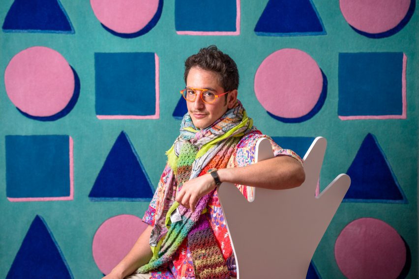 Photo if designer Adam Nathanial Furman wearing orange glasses and a green scarf against a geometric background