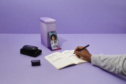 Photo of a hand scribbling in a notebook while in front of her a phone sat upright in a stand shows the face of a Black female doctor in a lab coat with a stethoscope around her neck. Medical gadgets also sit on the table beside the phone.