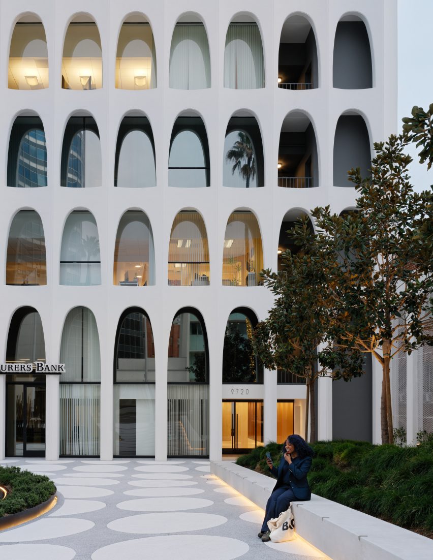 A person sitting on a white concrete bench in a plaza with an arched building behind it