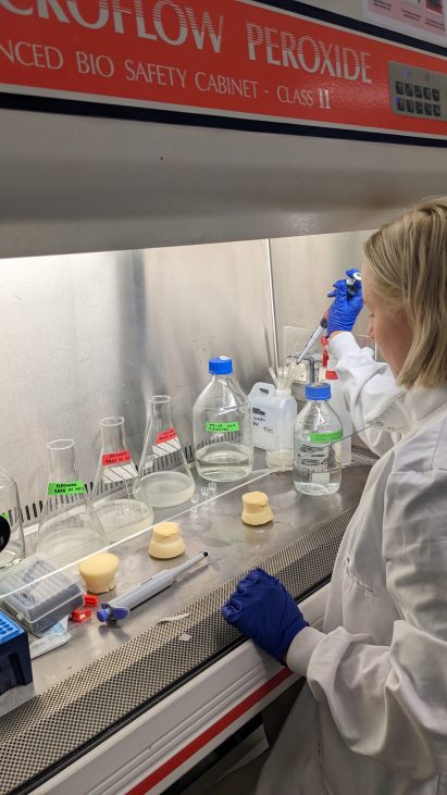 Photograph of a woman in a white lab coat using a pipette to move liquid between vessels