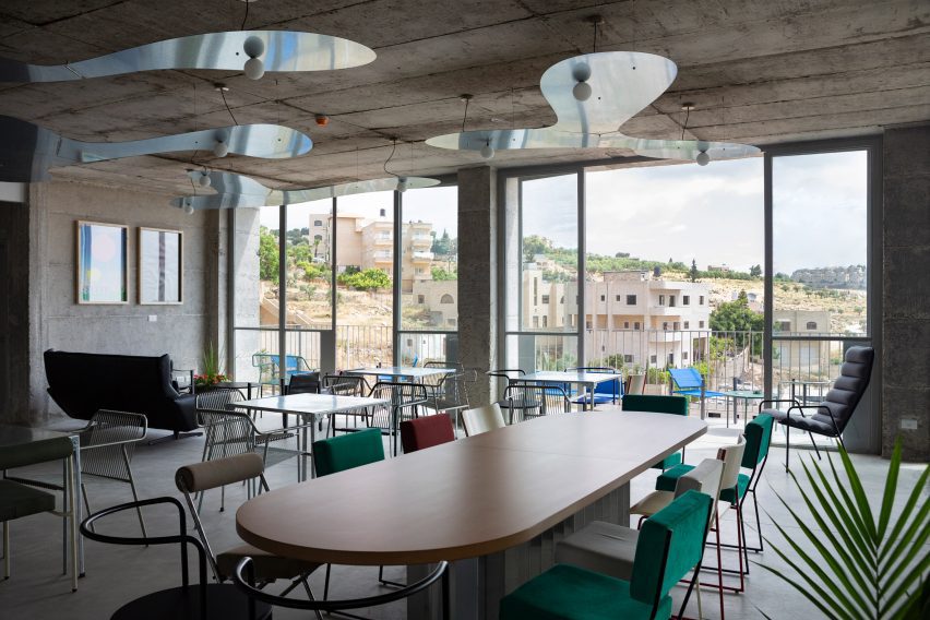 Photo of an open room in a building with concrete walls, floors and ceilings, with a large dining table and colourful mismatched chairs, a lounge area in one corner and views over a valley through floor-to-ceiling windows