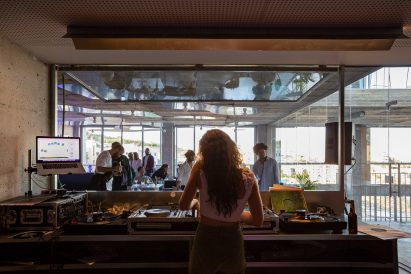 Photo of the inside of a DJ booth, looking out onto a cafe-like space where people mingle with drinks