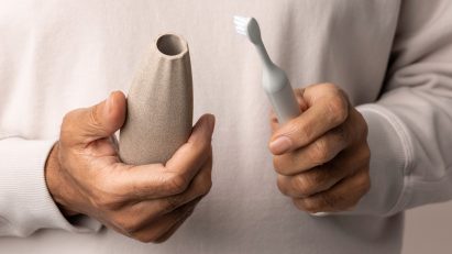 Close-up photo of a person's hands holding a toothbrush in one and a beige, ridges Access-ories grip in the other