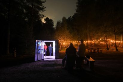 Photograph showing lit shipping container in woodland area at night