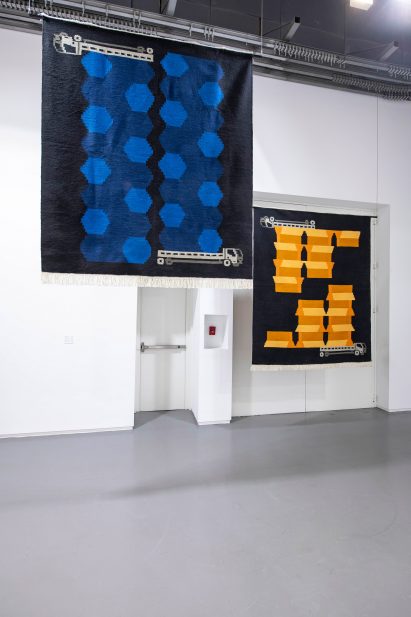 Photo showing two carpets hung up against a white wall decorated with colourful shapes and lorries