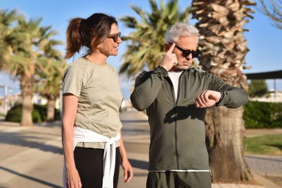 Photo of two middle-aged people in activewear paused on a footpath lined with palm trees. Both are wearing sunglasses and one is looking at the watch on his wrist