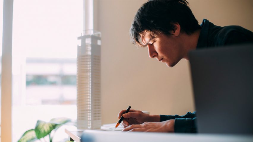 A person is writing next to a window, in front of which there is an architecture model