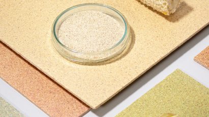 Photo of a flatlay of CornWall tiles by Circular Matters and StoneCycling in several muted, natural colours, arranged with a bare corn cob, a full corn cob with some of the husk on it and a small bowl of pale shredded organic material
