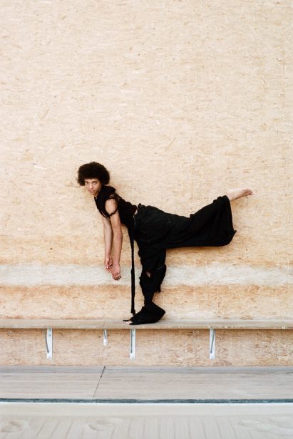 Standing model wears black garments and balances on a bench