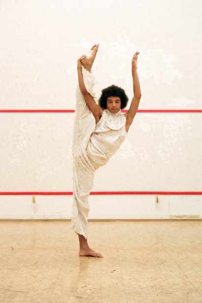 Model stands with one leg raised above their head, wearing a beige outfit