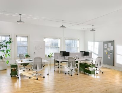 Two tables of modern desks with computer monitors are positioned in a light-filled room with light wood floors. 