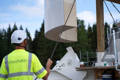 Photo of a worksite with a man in hi-vis operating machinery in the foreground and a large curved module being lowered into place in front of him