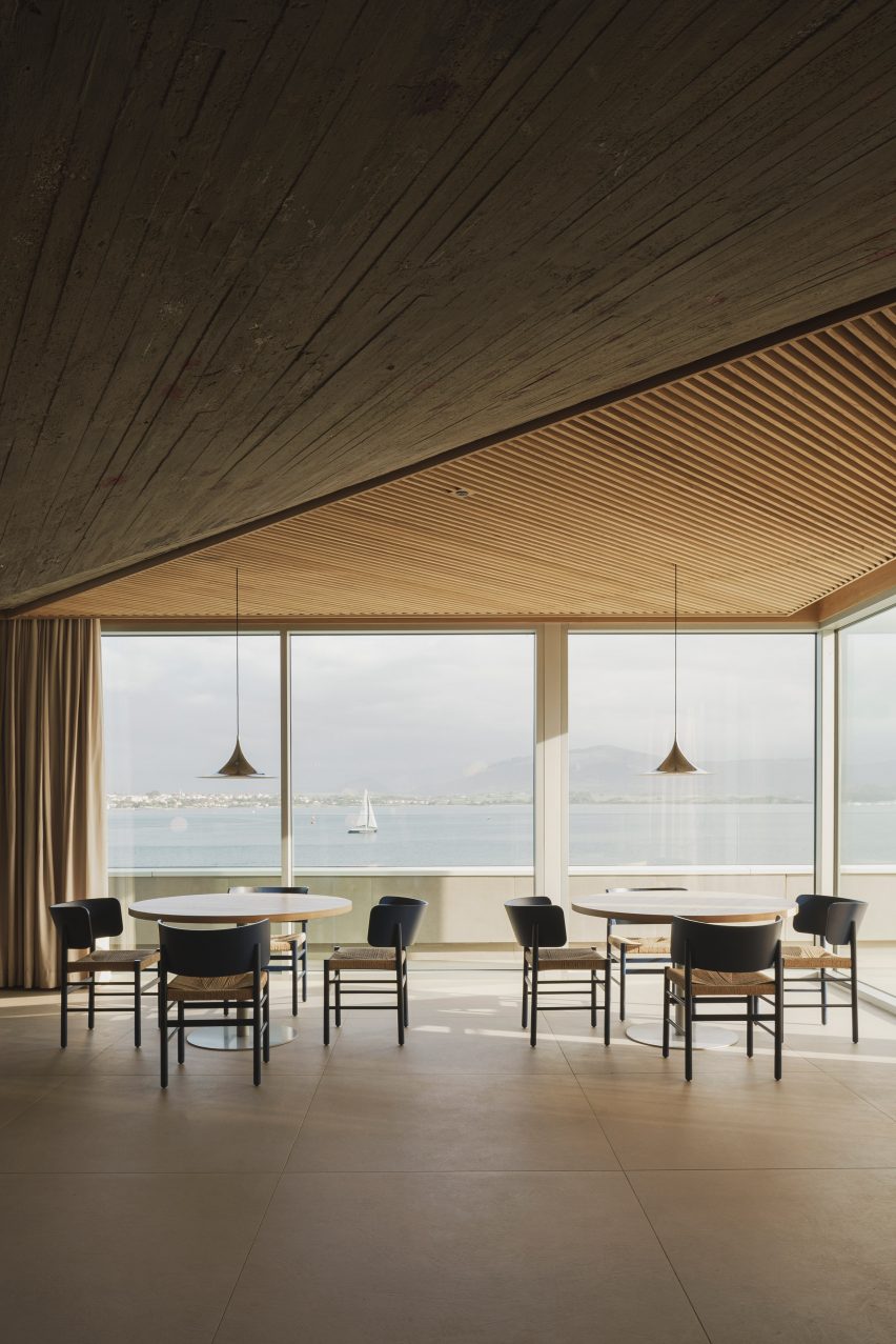 Dining tables overlooking views of the bay beneath a slatted timber ceiling inside a brutalist restaurant in Spain by Zooco Estudio