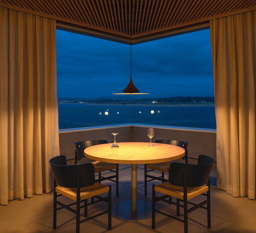Dining tables overlooking views of the bay by night from a frameless corner window inside a brutalist restaurant in Spain by Zooco Estudio