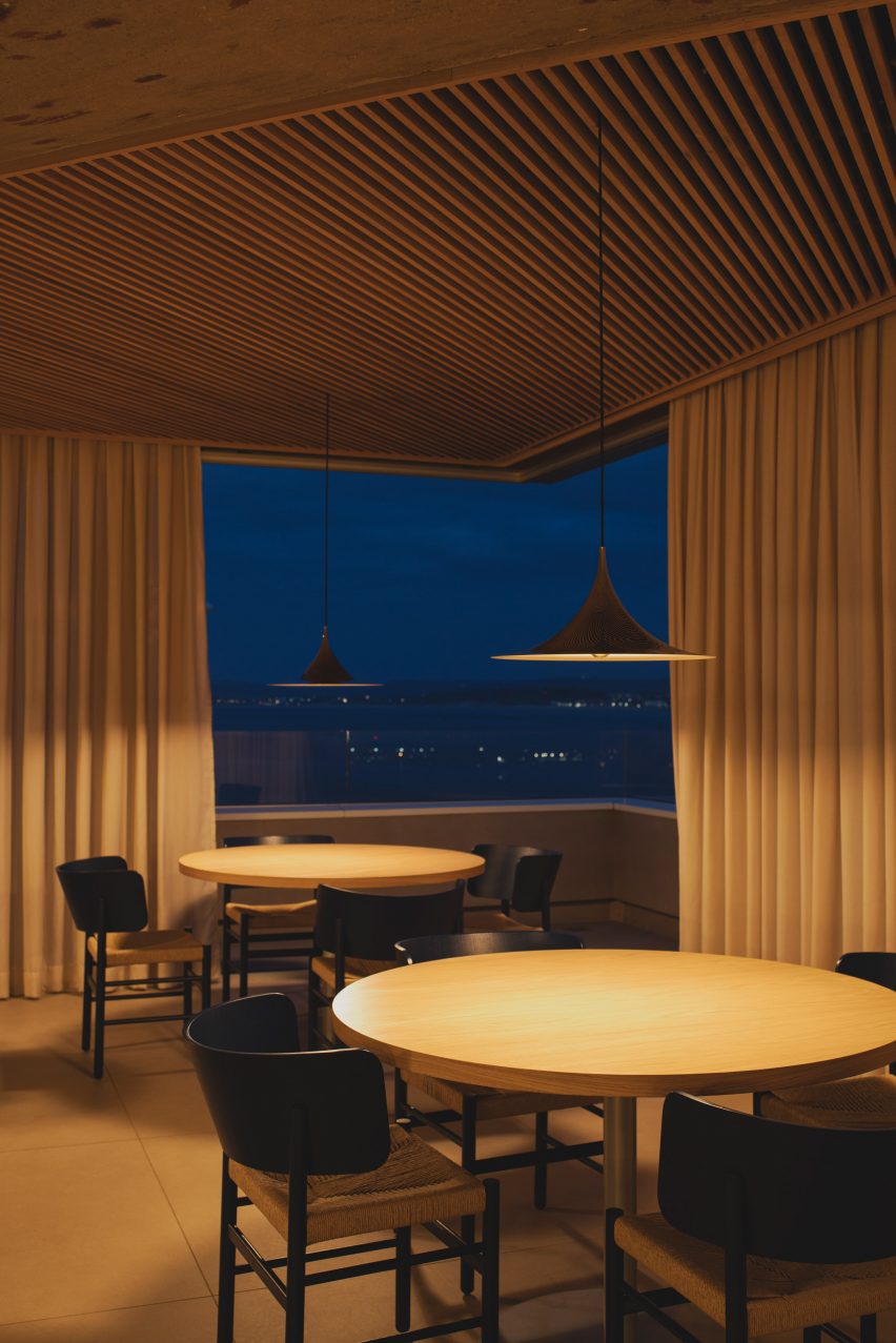 Dining tables overlooking views of the bay by night from a frameless corner window inside a brutalist restaurant in Spain by Zooco Estudio