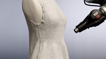 Still image of a white knit dress being formed to fit over a mannequin by a nearby robot arm hovering near it