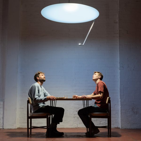 Photo of the Sunday Light creators Nat Martin and Sean Hammett sitting under their product, a bright disc suspended on the ceiling