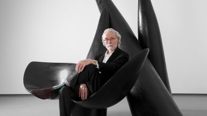 Photo of Wendell Castle in his later years sitting on one of his large, black, biomorphic sculptures
