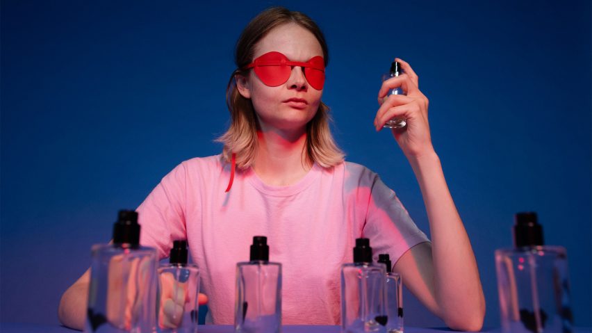 A photograph of a person holding a perfume bottle to their face. They are wearing a pink t-shirt and a red blindfold over their face, against a blue backdrop.