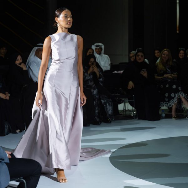 A person walking on a white runway floor with people in chairs around them. They are wearing a silver dress.