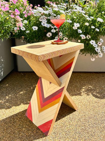 A triangular shaped wooden side table with geometric patterns in red, yellow and brown tones, placed on gravel.