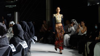 A person walking on a white runway floor with a black backdrop, wearing a red skirt and a yellow top with blue sleeves.